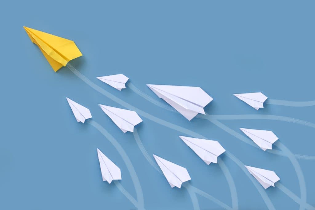 Conceptual image of a yellow paper airplane leading a group of white paper airplanes on a blue background, symbolizing leadership, innovation, and standing out from the crowd