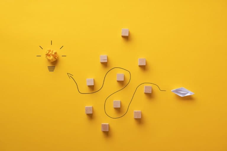 Creative concept of problem-solving with a crumpled yellow paper lightbulb, wooden blocks, and a winding path leading to a paper boat on a bright yellow background.