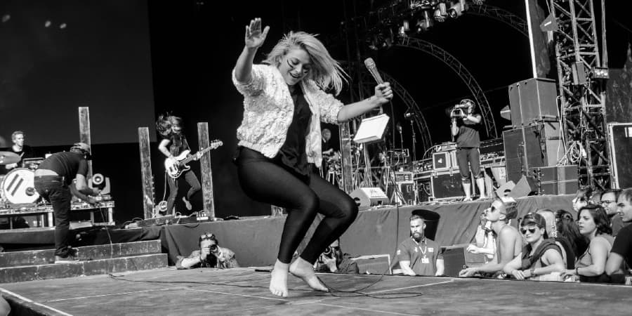 Black and white photo of a barefoot female performer energetically singing on stage during a live concert, surrounded by band members and an engaged audience
